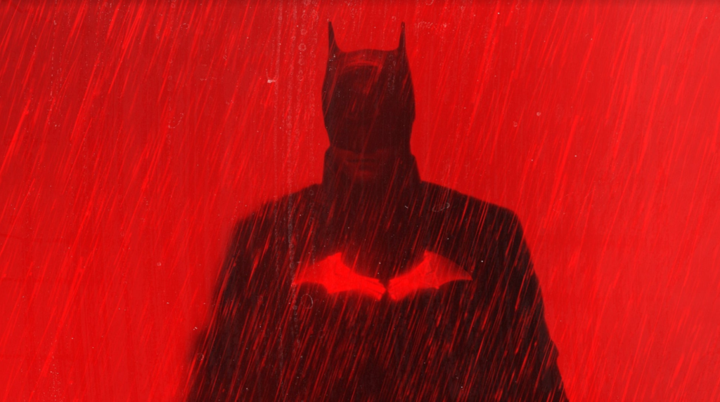 The Batman stands in the rain as red light from a vehicle shines on.

 