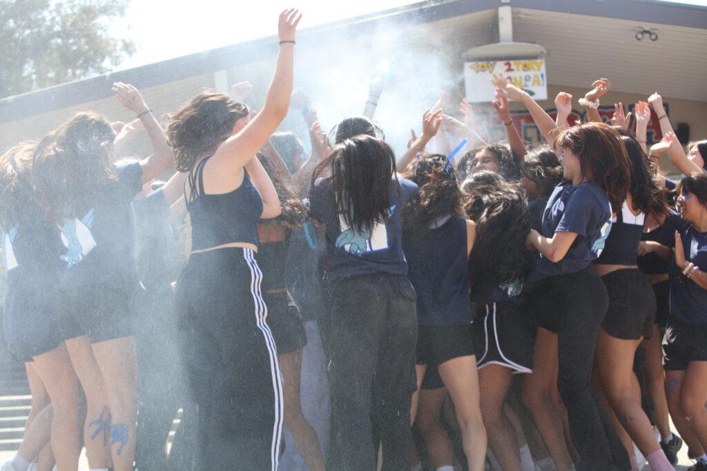 Class of 2025 performers crowd together and cheer under a cloud of blue powder at the end of their performance.