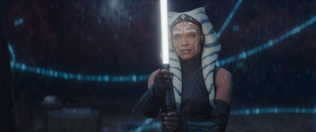 Ahsoka holds up her white lightsaber with a large celestial map projected in the background.