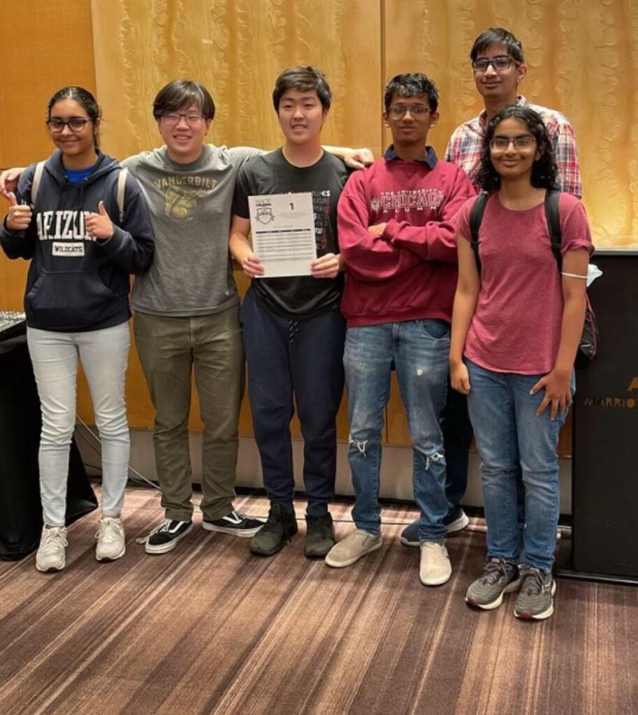 Senior Simarya Ahuja and Class of ‘23 alumni Adam Xu, Anthony Wang, Siddharth Kamannavar, Nithya Krishna and Nilay Mishra (left to right) hold up the schedule card of the team they beat on May 27, 2023, the first day of prelims at Nationals.