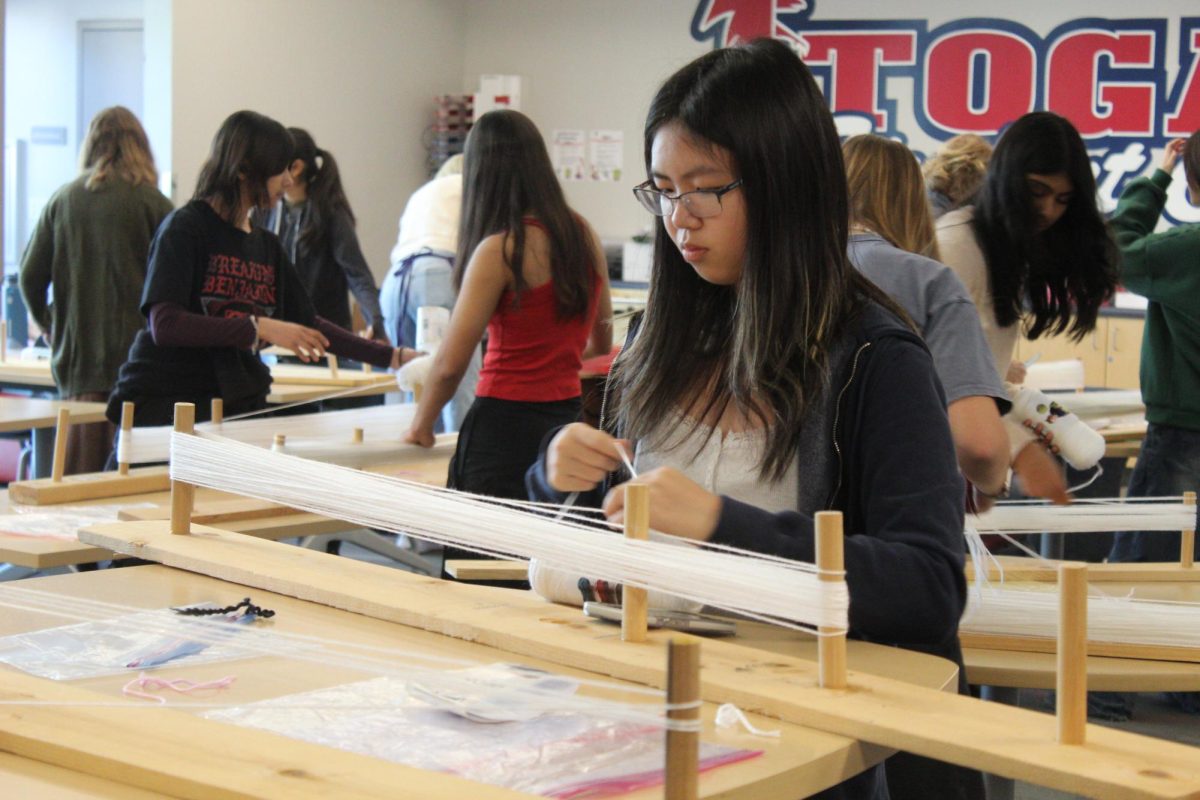 Senior Lily Jiang helps out with the Magic Yarn station, threading yarn into a loom.